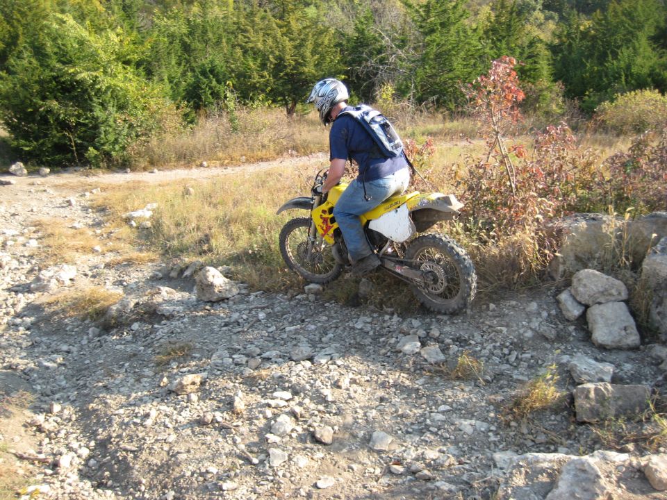 Rider on yellow dirt bike navigating a rocky, off-road trail with trees in the background