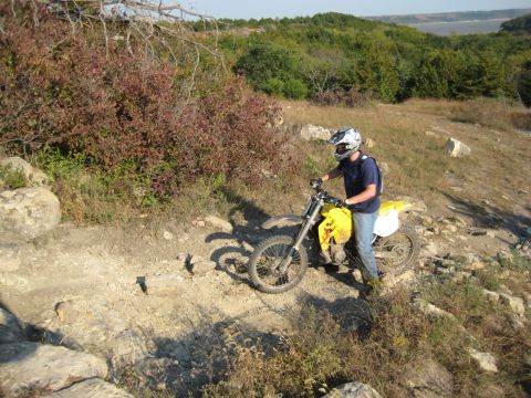 1993 Suzuki RMX250 - Rider on a yellow dirt bike navigates a challenging rocky trail with a scenic overlook in the background.