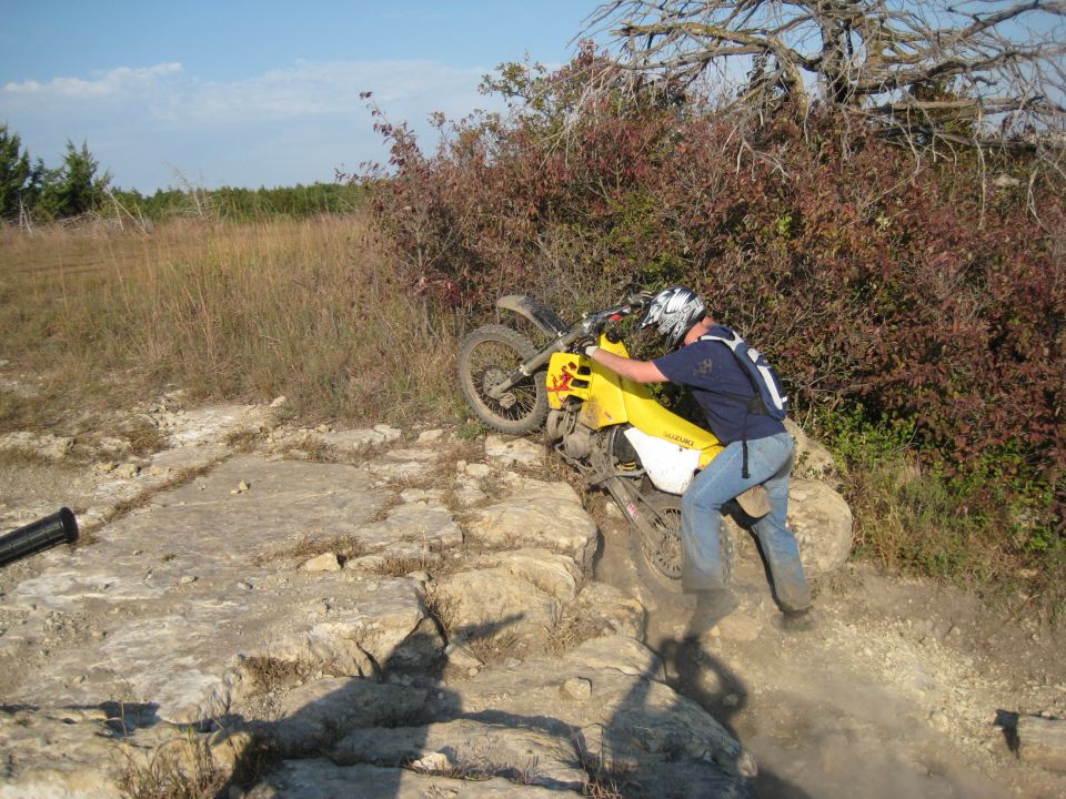 Guy struggling to push a yellow dirt bike up a rocky trail. He's wearing jeans and a helmet
