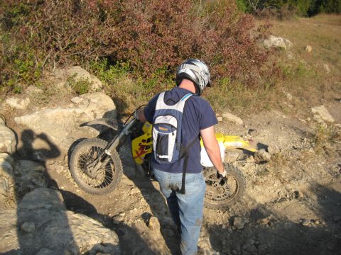 1993 Suzuki RMX250 - A rider walks a yellow dirt bike through a rocky, off-road trail.