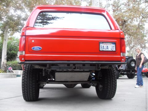 1990 Ford Bronco - Rear view of a red 1990 Ford Bronco, showing its modified bumper and suspension. Another vehicle is parked nearby.