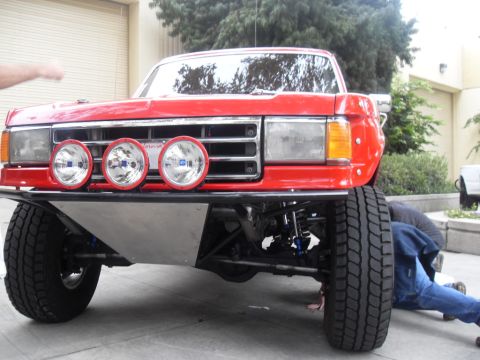 1990 Ford Bronco - A person works on the suspension of a red off-road truck with auxiliary lights mounted on the front.