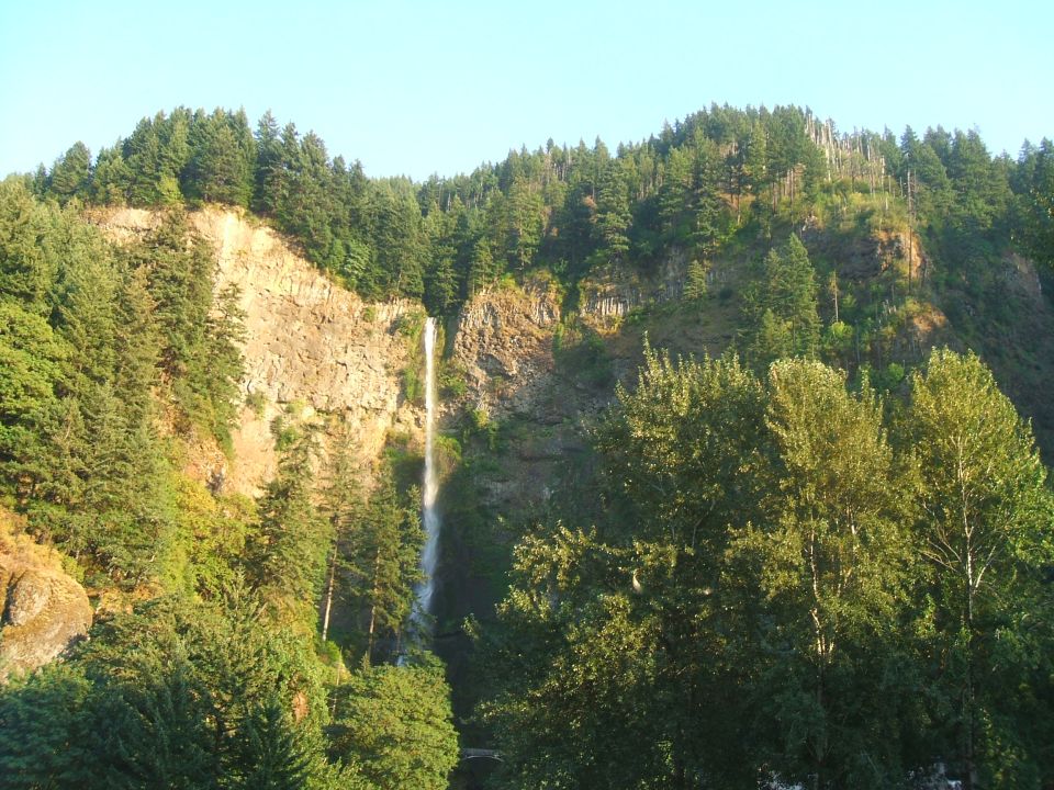 Waterfall cascading down a rocky cliff face surrounded by lush green trees under a clear blue sky