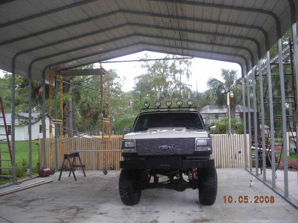 Lifted Ford truck with roof lights and aftermarket bumper parked under a metal carport