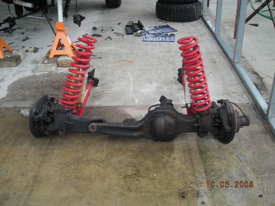 Front axle assembly with red coil springs and brake components, resting on concrete