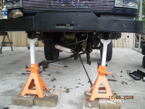 1993 Ford Bronco - The front suspension of a dark SUV is elevated on jack stands, with various tools visible on the ground around it.