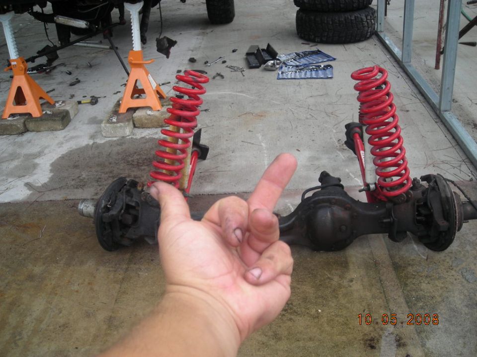 Front axle assembly with red coil springs and shock absorbers, partially obscured by a hand making a peace sign. Tools and jack stands are visible in the background