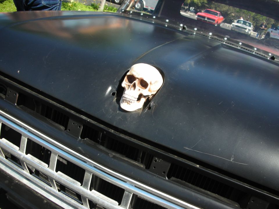 Black vehicle hood with skull ornament and chrome grille. Other cars visible in reflection