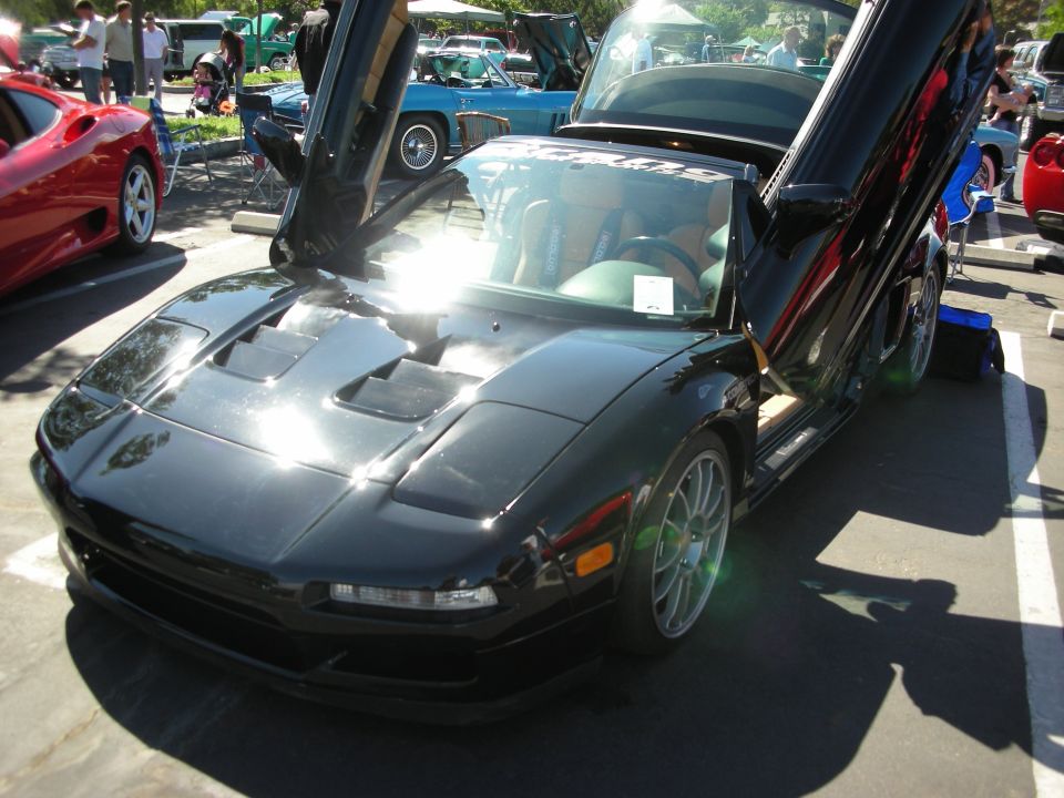 Black sports car with scissor doors open, parked at a car show. Other cars visible in background