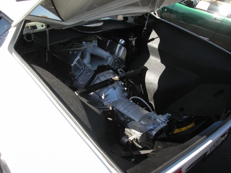 White car's engine compartment with a supercharger and transmission visible. Green car in background