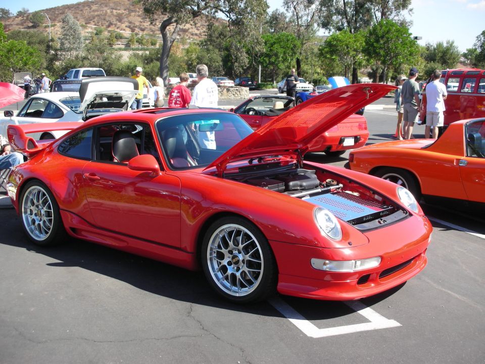 Red Porsche 911 with hood open, showing engine bay. Other cars and people are visible in the background at a car show