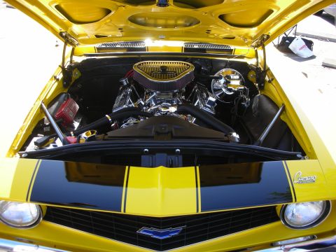 1998 Ford Crown Victoria - The engine bay of a yellow 1969 Chevrolet Camaro is open, revealing a chrome engine and black racing stripes.