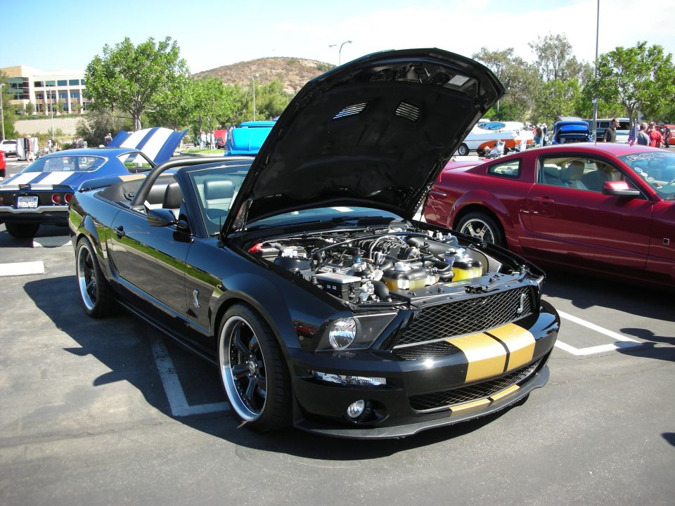 Black convertible Mustang with gold stripes on the hood and grille, engine exposed. Red Mustang parked to the right, blue and white car to the left