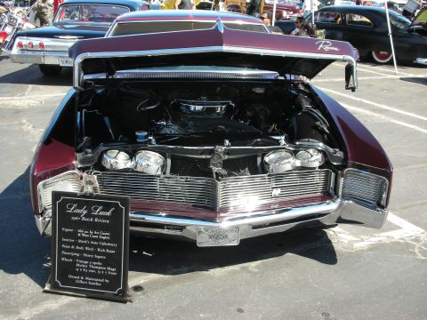 1998 Ford Crown Victoria - A burgundy 1967 Buick Riviera is displayed with its hood open, revealing the engine and a descriptive sign at a car show.