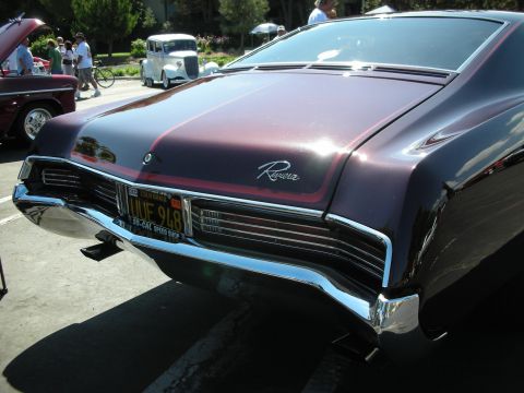 1998 Ford Crown Victoria - A classic maroon Buick Riviera is parked at a car show, with other vehicles and people in the background.