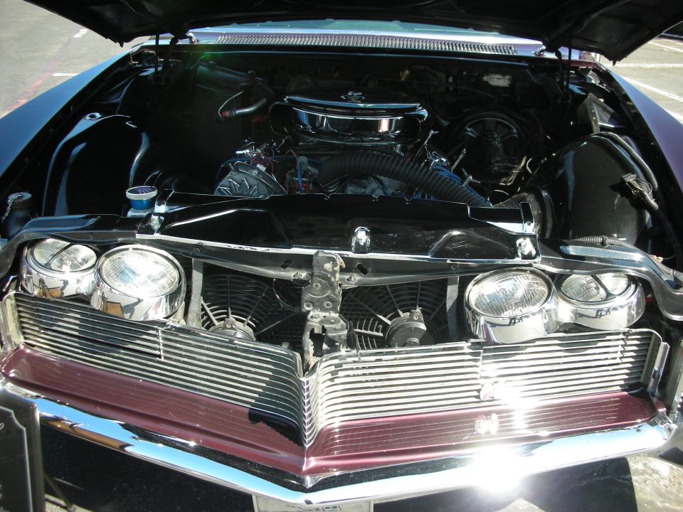 Engine bay of a classic car with a large chrome grille and dual headlights. Hood is open, showing the engine and fans