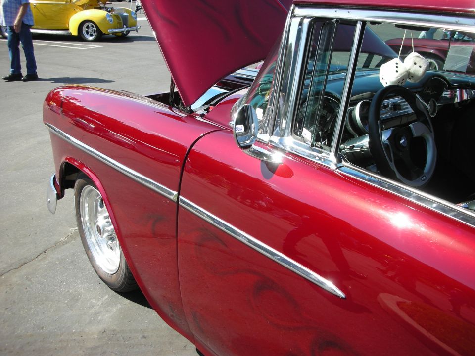 Red classic car interior showing steering wheel and fuzzy dice hanging from rearview mirror. Another yellow car is visible in the background