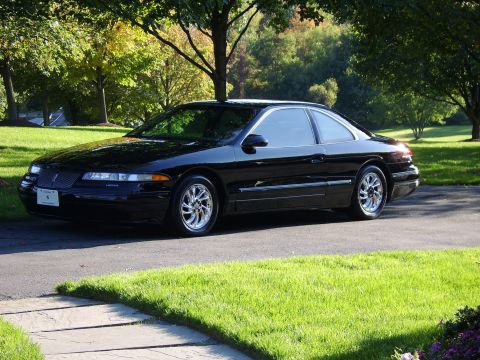 1996 Mercury Grand Marquis - A black coupe with distinctive chrome wheels is parked on a paved driveway surrounded by lush green grass and trees.