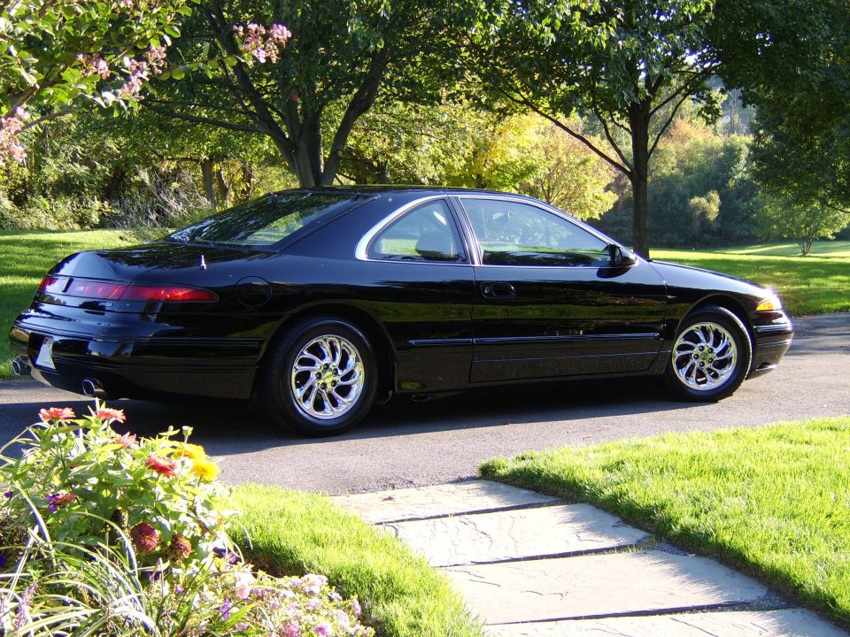 Black coupe parked on driveway, rear passenger-side view, chrome wheels, in garden setting
