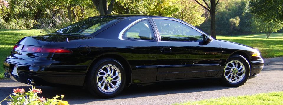 Black coupe viewed from rear side, chrome wheels, on driveway with trees behind