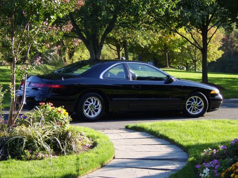 1996 Mercury Grand Marquis - A black coupe with chrome wheels is parked on a paved driveway surrounded by lush green grass and trees.
