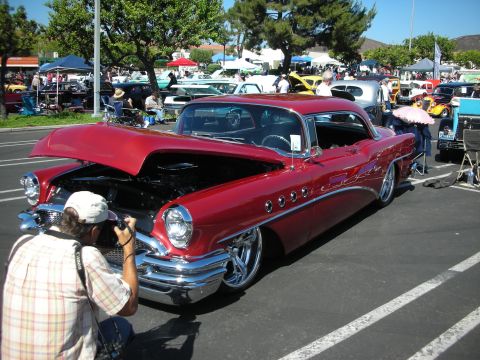 1998 Ford Crown Victoria - A person photographs a shiny red vintage car at an outdoor car show with other vehicles in the background.