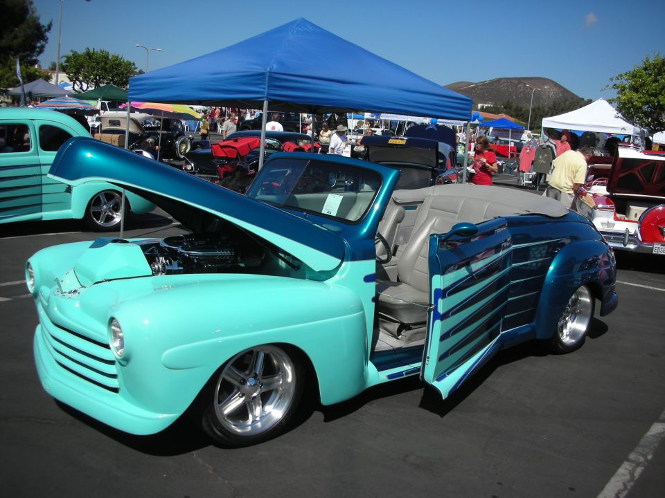 Teal and blue convertible hot rod with hood and door open, showing engine and interior. Other cars and tents in background