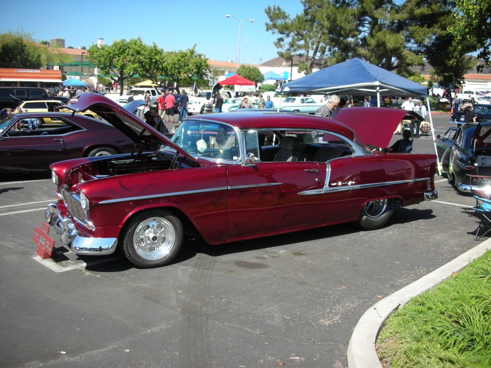 Red 1955 Chevrolet Bel Air coupe with hood open, parked at a car show. Another car is visible to the left