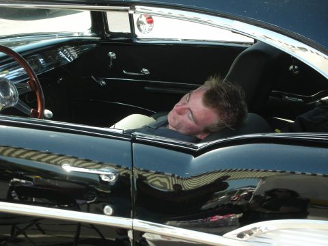 1998 Ford Crown Victoria - A man takes a nap inside a classic car, its interior a distinctive steering wheel and chrome trim.