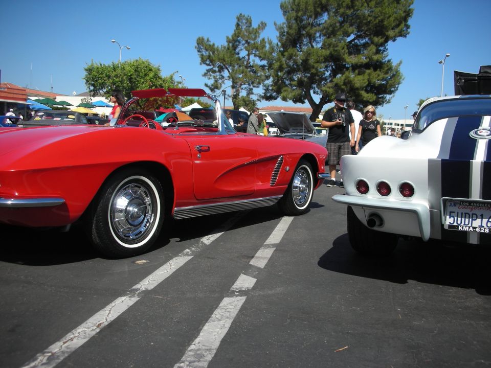 Red convertible sports car side view, white wall tires, chrome accents. Another car with blue stripes visible
