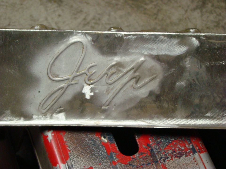Close-up of a chrome Jeep emblem on a metal surface, with some scuff marks and a red and black object below
