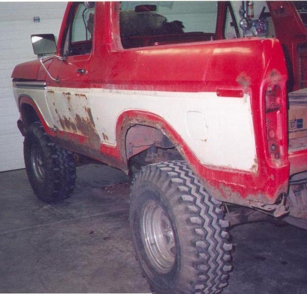 Rear three-quarter view of a lifted red and white Ford Bronco with rust and large off-road tires in a garage