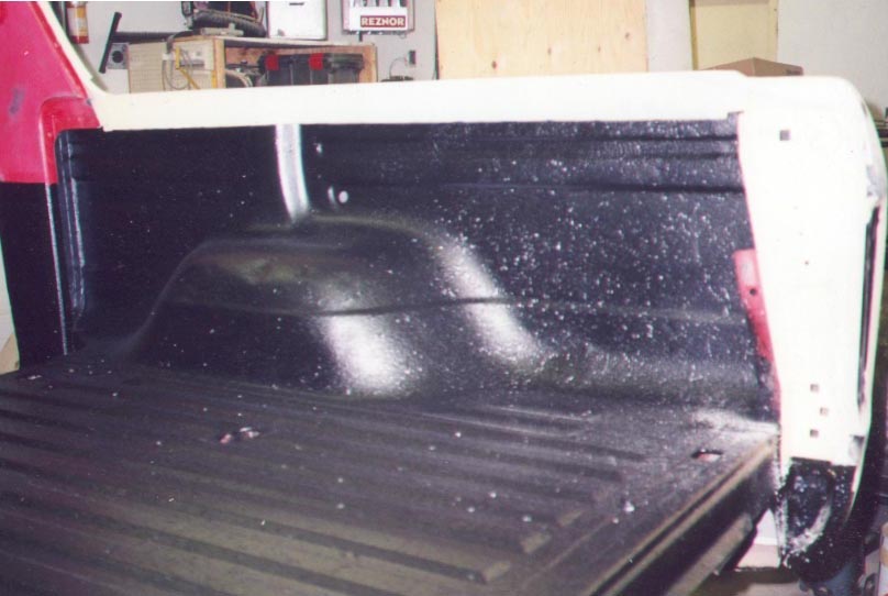 Truck bed interior with glossy black spray-on bedliner over ribbed floor and wheel well, in a shop setting