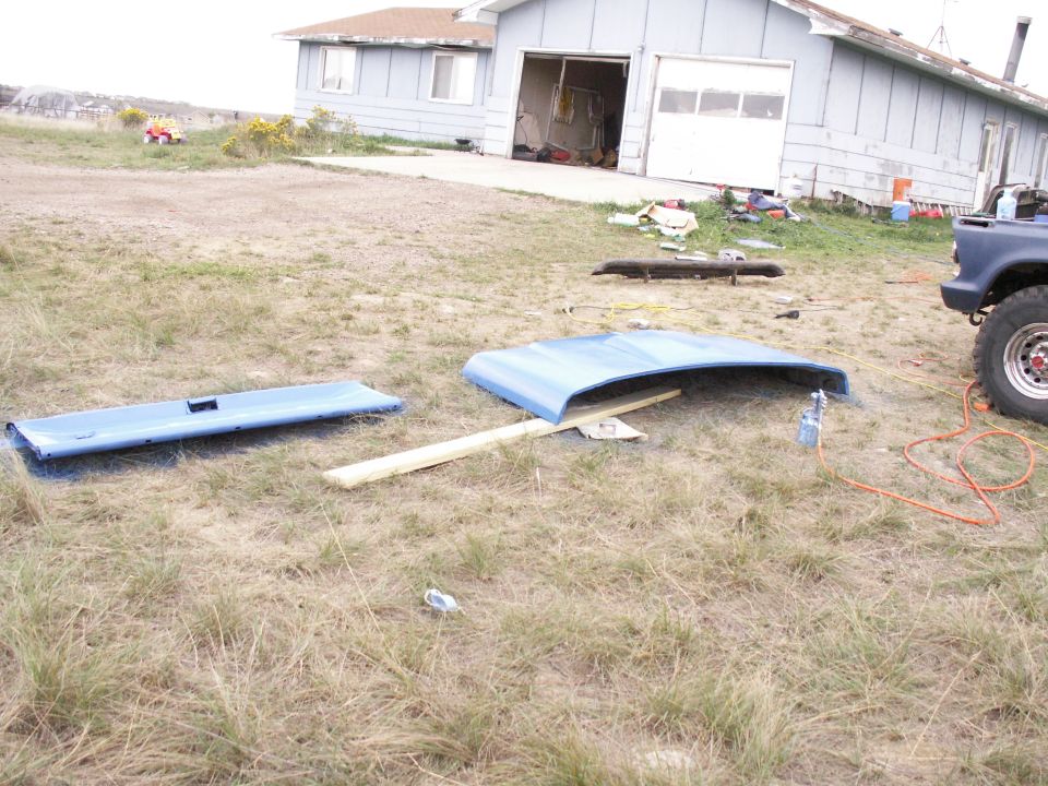 Blue car hood and tailgate lying on dry grass near a house and a blue truck