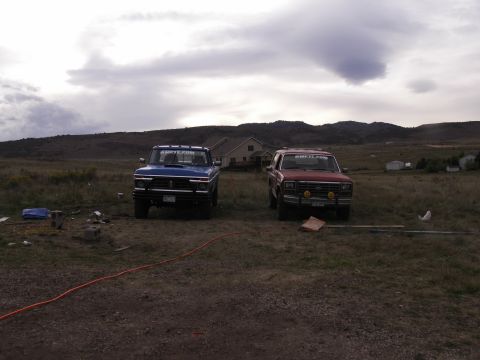 1977 Ford F-250 - Two trucks are parked in a grassy area with a house and hills in the background under a cloudy sky.