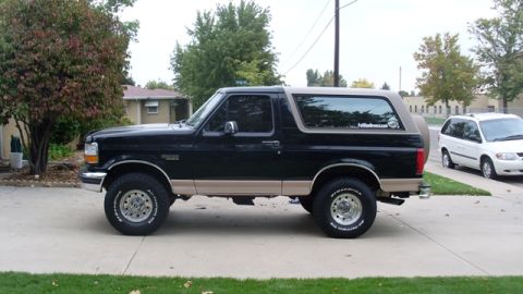 1996 Ford Bronco - A black and tan 1996 Ford Bronco is parked on a driveway next to a white minivan.