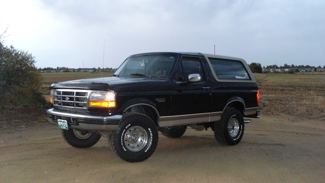 Lifted black and tan Ford Bronco on a dirt lot, front 3/4 view, overcast sky