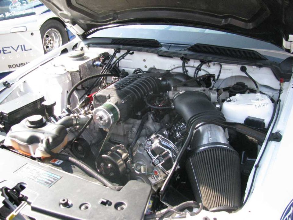 Supercharged V8 engine bay with custom intake and polished components. Another white car with 'DEVIL SPORTS ROUSH' visible is parked nearby