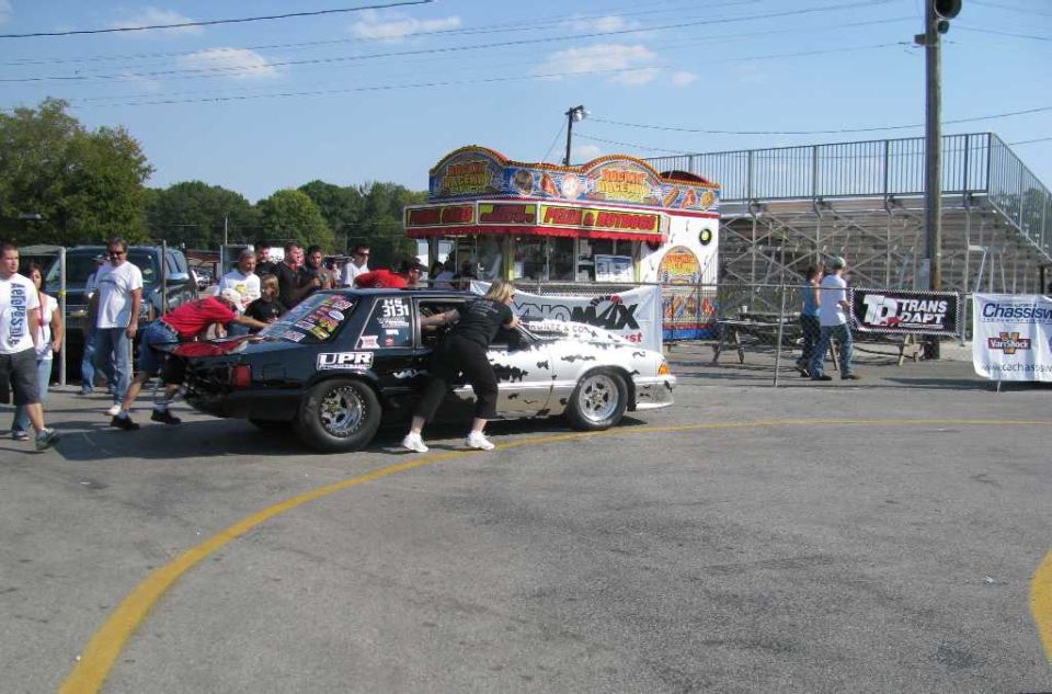 Two drag racing cars, one black and one white, are being pushed by people on a track. A food stand and bleachers are in the background