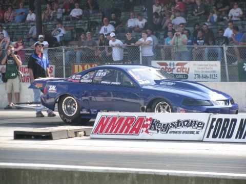 2003 Mercury Marauder - A blue drag car with racing decals is positioned on a trailer at a drag strip, with spectators watching from the stands.