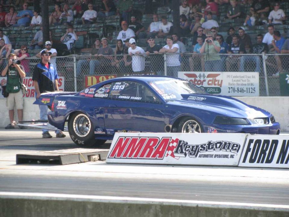 Blue drag car with racing decals on a trailer, spectators in bleachers