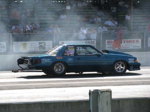2003 Mercury Marauder - A teal drag car with numerous racing decals and a prominent hood scoop sits on a drag strip, emitting smoke.