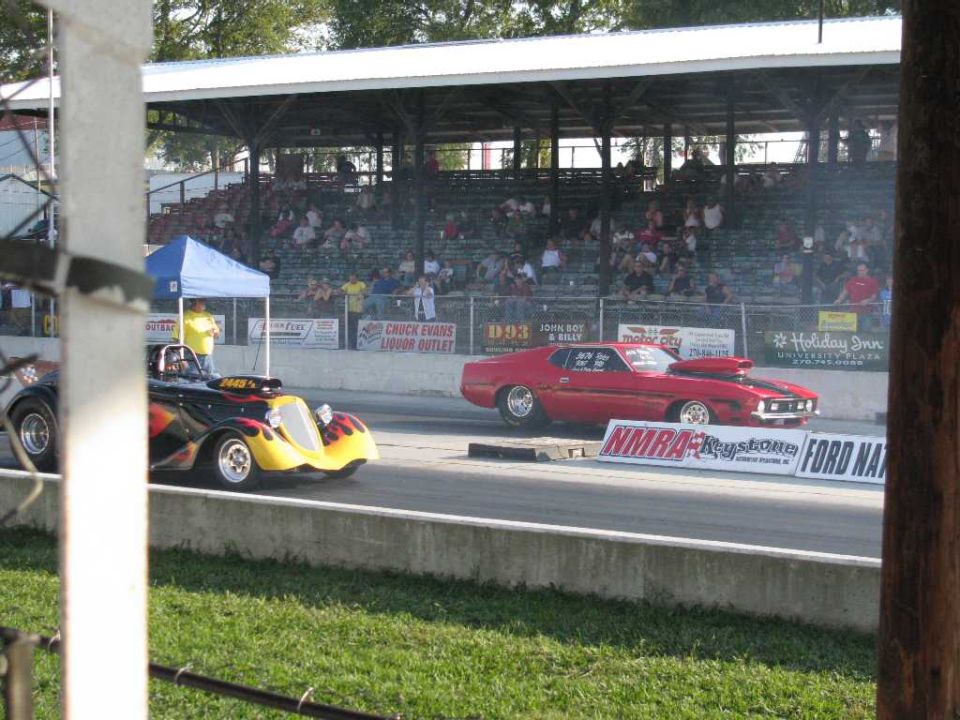 Two drag cars racing down a track, one red and one yellow/black with flames. Spectators in the stands