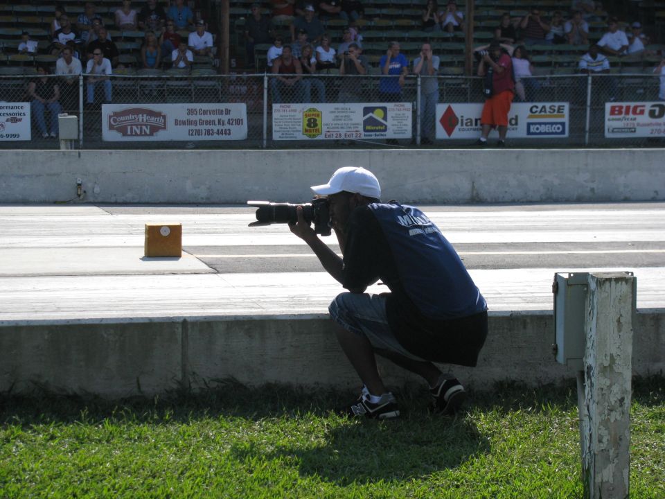 Photographer crouches by a concrete barrier at a drag strip, aiming a camera down the track