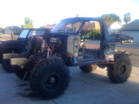 1994 Ford Bronco - This heavily modified truck chassis, stripped down and equipped with oversized tires, sits in a driveway with another vehicle visible in th…