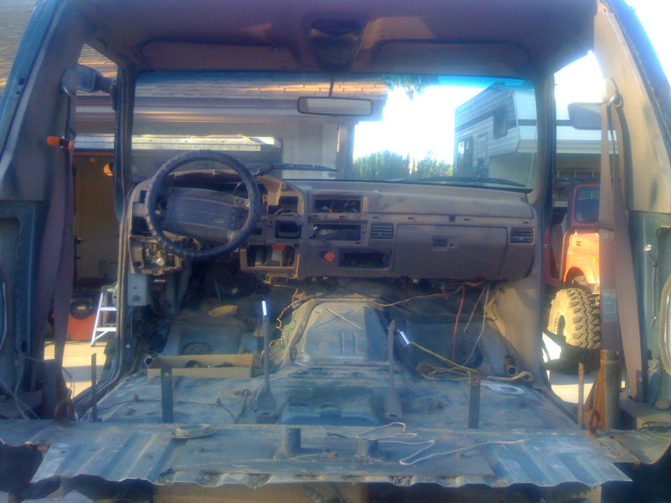 SUV interior stripped bare, showing dashboard, steering wheel, and exposed metal floor. A trailer and another vehicle are visible through the open doors