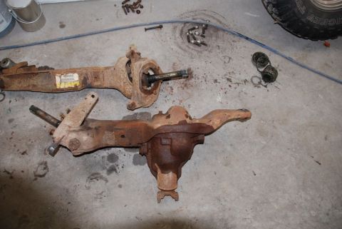 1996 Ford Bronco - Two heavily rusted front axle assemblies are laid out on a concrete floor, with various bolts scattered nearby.