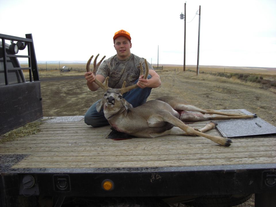 Hunter holding buck antlers on a wood-deck flatbed truck bed in open prairie