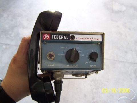 1998 Ford Crown Victoria - Hand holding a vintage Federal Signal 'Interceptor' siren control head with a black PA handset clipped to its side. The blue faceplate has…