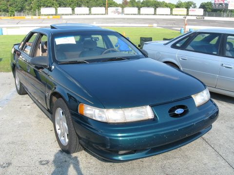 1993 Ford Taurus - A dark green sedan with a sunroof is parked next to a silver sedan at an outdoor venue.